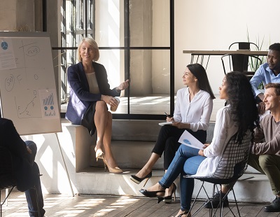Happy older female mentor and diverse business team discuss presentation in modern office, middle aged mature businesswoman leader coach training young multiracial workers group at corporate meeting