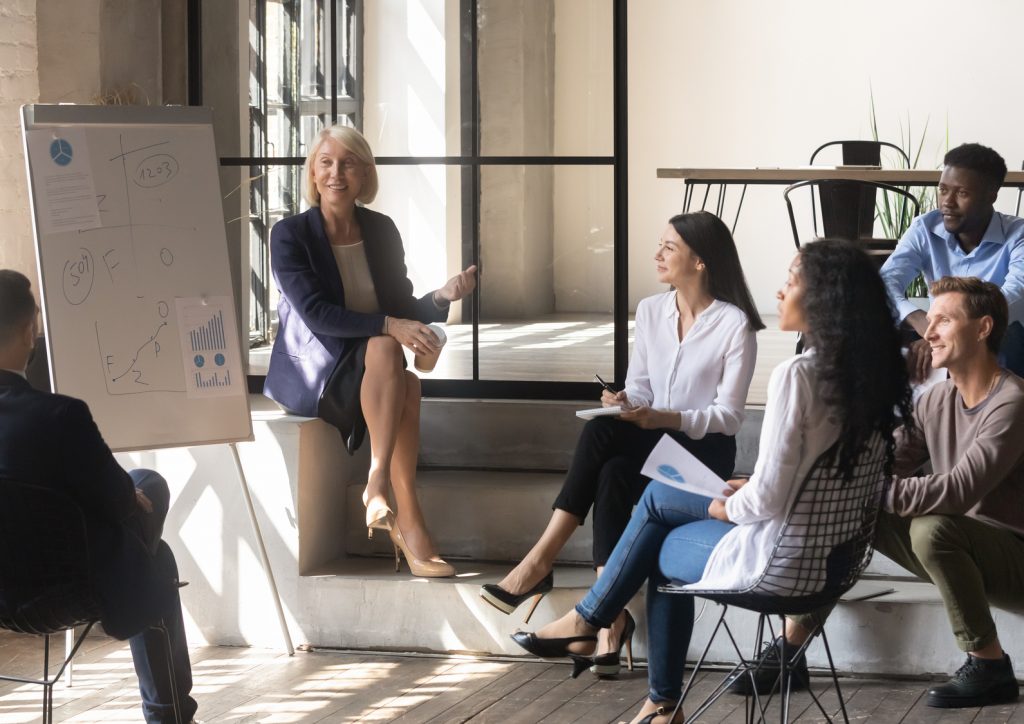 Happy older female mentor and diverse business team discuss presentation in modern office, middle aged mature businesswoman leader coach training young multiracial workers group at corporate meeting